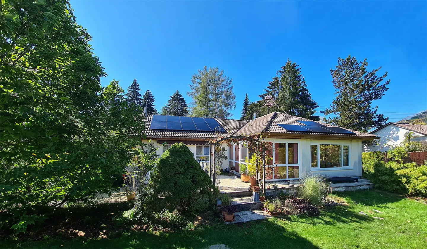 Ein Einfamilienhaus mit Photovoltaikanlage auf dem Dach, umgeben von einem grünen Garten mit Bäumen, Sträuchern und einer überdachten Terrasse. Der Himmel ist blau und die Solarmodule sind gut sichtbar auf den Dachflächen installiert.