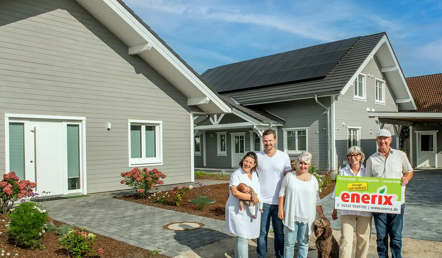 Eine glückliche Familie steht vor einem modernen Haus mit einer installierten Photovoltaikanlage auf dem Dach. Im Vordergrund hält ein älteres Paar ein Schild mit dem enerix-Logo und der Aufschrift "Photovoltaik · Stromspeicher · Ladetechnik – Energie zum Nulltarif". Der Garten ist gepflegt, und die Szene strahlt Nachhaltigkeit und Zufriedenheit aus.