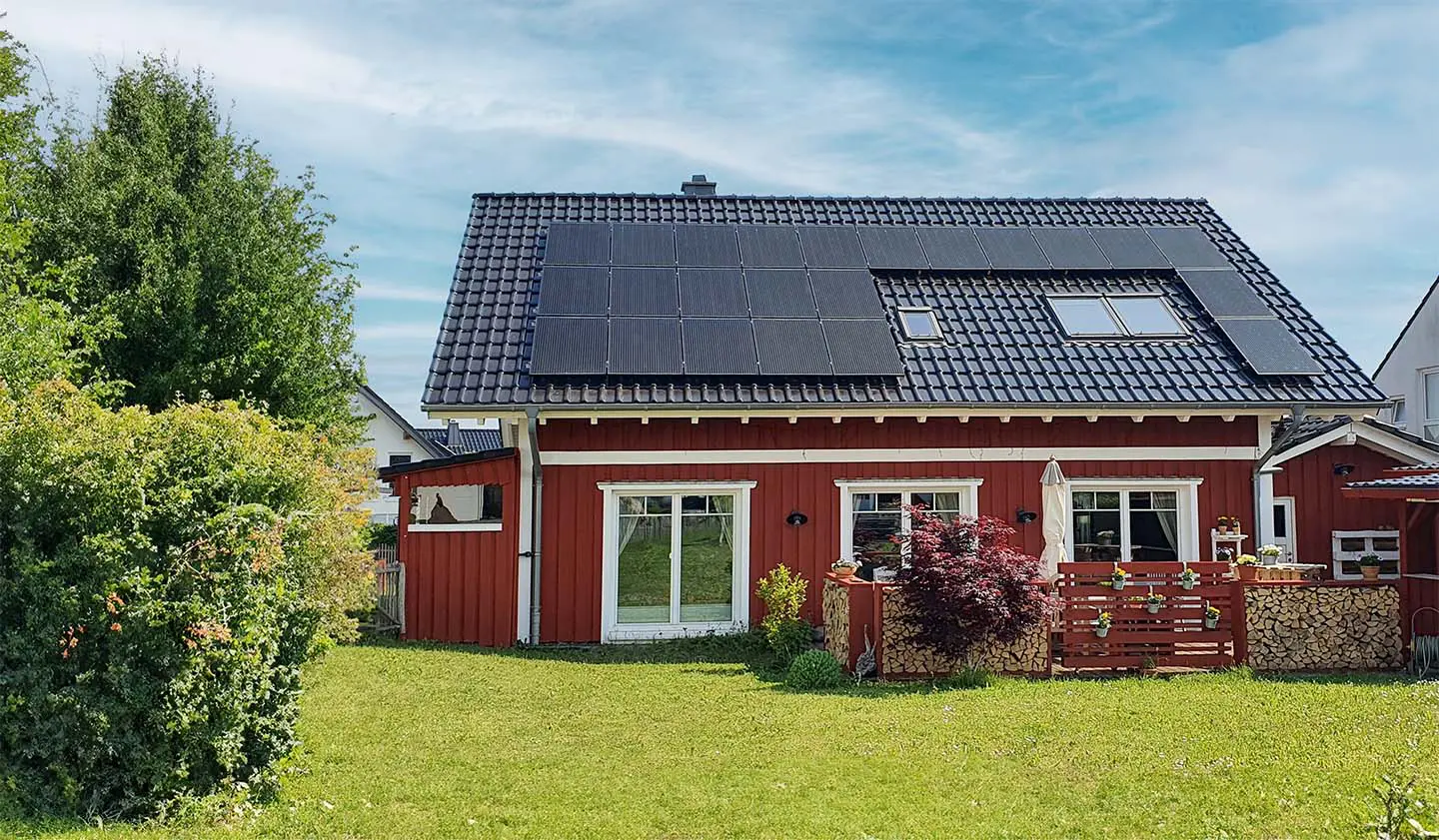 Ein modernes rotes Einfamilienhaus mit schwarzem Dach, auf dem eine Photovoltaikanlage installiert ist. Umgeben von grünem Rasen und Bäumen, vermittelt das Bild Nachhaltigkeit und die Nutzung erneuerbarer Energien.