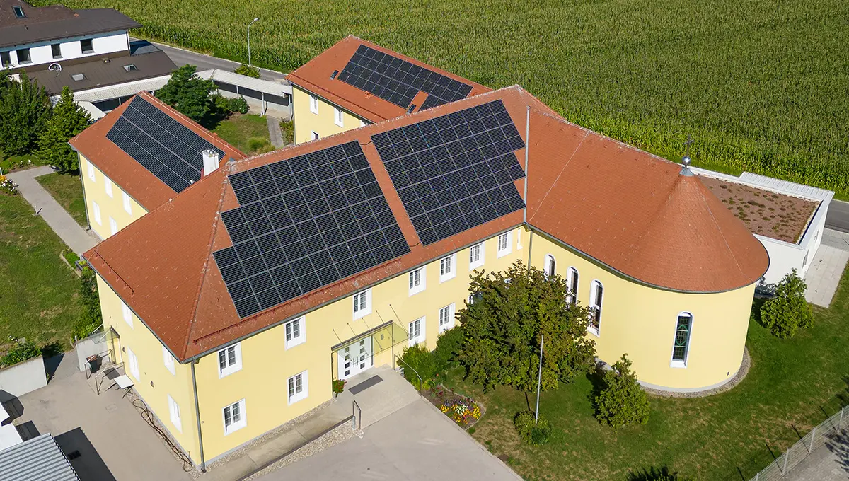 Ein großes, gelbes Kloster mit einem roten Ziegeldach, das von Solarpaneelen bedeckt ist. Das Gebäude hat eine rechteckige Form mit einem abgerundeten Anbau auf der rechten Seite. Es ist von grünen Rasenflächen, Bäumen und Blumenbeeten umgeben, während im Hintergrund ein großes Feld zu sehen ist. Die Aufnahme wurde aus der Vogelperspektive gemacht und zeigt auch benachbarte Gebäude.