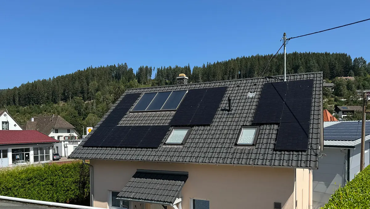 Ein Einfamilienhaus mit einem geneigten Ziegeldach, das mit mehreren Solarpaneelen ausgestattet ist. Im Hintergrund sind dichte Nadelwälder und ein klarer blauer Himmel zu sehen. Das Haus befindet sich in einer ruhigen, ländlichen Umgebung, die von weiteren Häusern und Gebäuden umgeben ist.