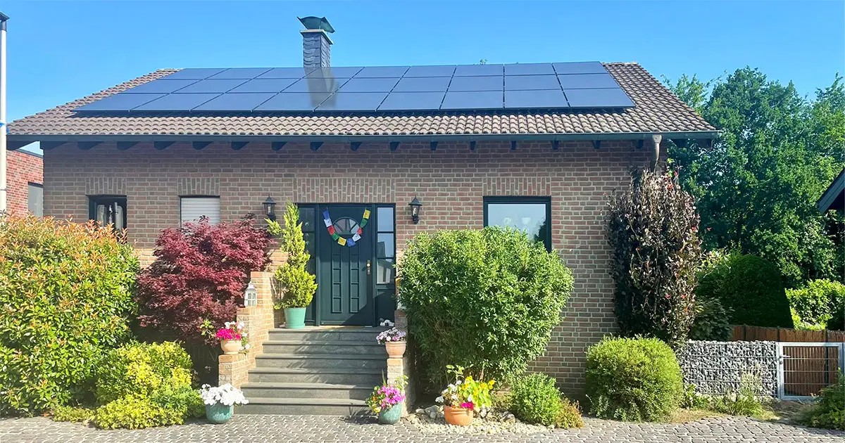 Das Bild zeigt ein Einfamilienhaus mit einer Photovoltaikanlage auf dem Satteldach. Die schwarzen Solarmodule sind gleichmäßig verteilt. Das Haus hat eine Backsteinfassade und ist von gepflegten Pflanzen und Blumen umgeben.