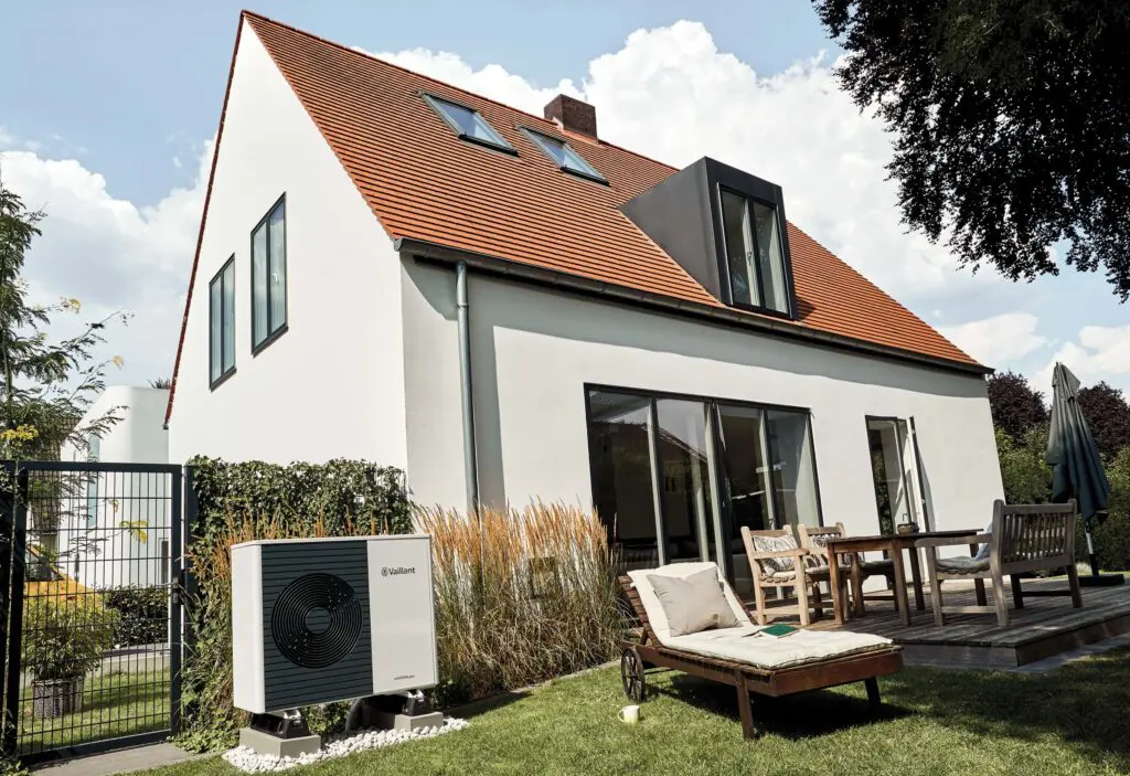 Modernes Einfamilienhaus mit rotem Ziegeldach und großer Terrasse. Im Vordergrund steht eine Vaillant-Luft-Wasser-Wärmepumpe im Garten.