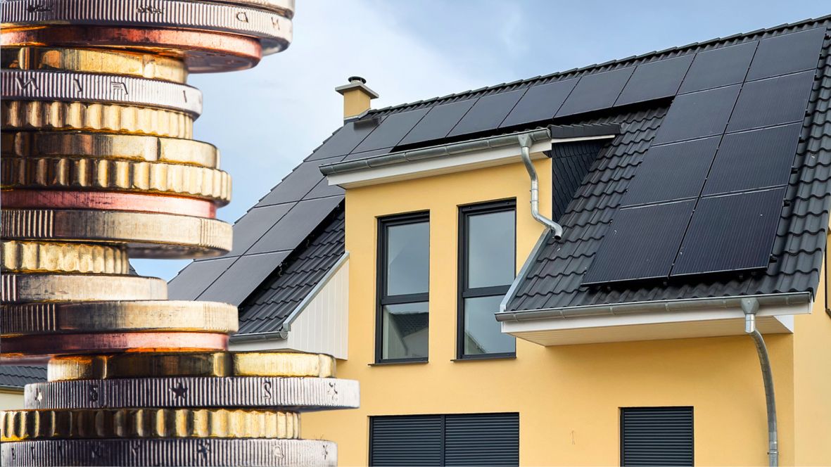 Ein gelbes Haus mit einem schwarzen Dach auf dem eine schwarze Solaranlage installiert ist. Im Vordergrund sind einige gestapelte Euro-Münzen zu sehen.