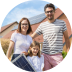 Eine Famile mit einem kleinen Solarmodul in der Hand vor einem Haus mit Photovoltaikanlage auf dem Dach.