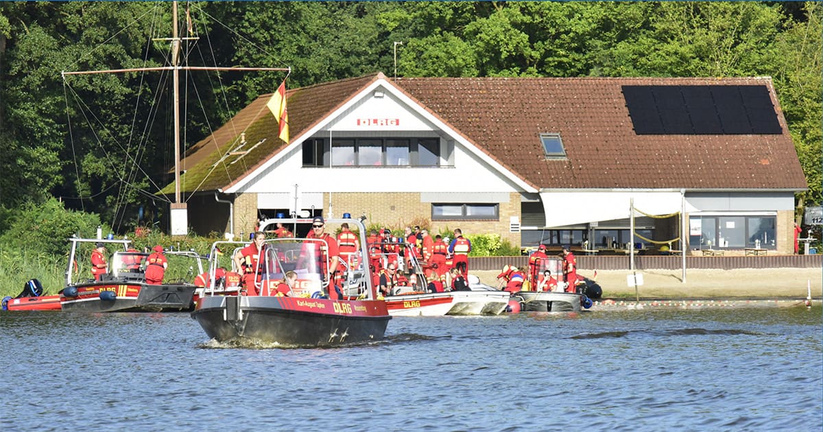 Das Bild zeigt eine DLRG-Station (Deutsche Lebens-Rettungs-Gesellschaft) am Ufer eines Sees, umgeben von zahlreichen Rettungsbooten und DLRG-Mitgliedern in roten Uniformen. Auf dem Dach des Gebäudes ist eine Photovoltaikanlage mit mehreren Solarmodulen installiert. Im Vordergrund ist ein Rettungsboot auf dem Wasser zu sehen, das von mehreren DLRG-Mitgliedern besetzt ist. Das Gebäude ist von Bäumen umgeben, und der Bereich wirkt gut besucht und belebt.