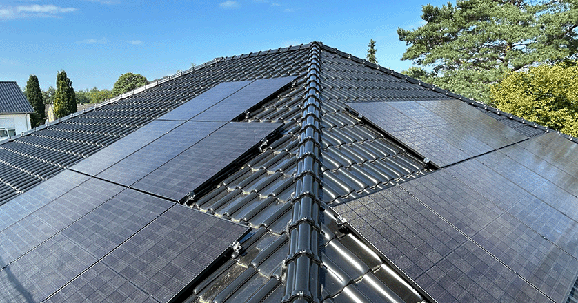 ein Hausdach mit schwarzes Ziegeln und einigen Solarmodulen um Hintergrund sind Himmel und Bäume zu sehen.