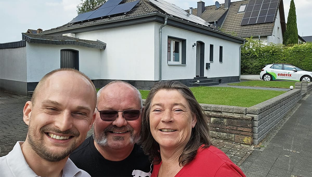 Das Bild zeigt zwei zufriedene Kunden, ein Mann und eine Frau, gemeinsam mit einem Mitarbeiter von enerix vor ihrem Einfamilienhaus, auf dessen Dach eine Photovoltaikanlage installiert ist. Im Hintergrund steht ein Firmenfahrzeug von enerix