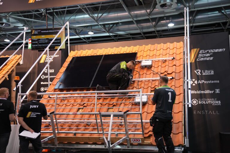 Teilnehmer bei den European Solar Games montieren Solarmodule und Wechselrichter auf einem Modellhausdach unter Beobachtung einer Jury – ein Wettbewerb zur Förderung von Fachkompetenz im Bereich Photovoltaik-Installation.