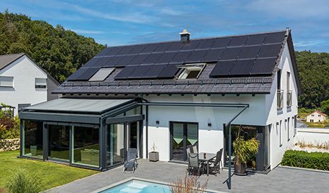 Modernes Einfamilienhaus mit großer Photovoltaikanlage auf dem Dach, kombiniert mit einem Wintergarten und einer Terrasse mit Pool. Die Anlage symbolisiert nachhaltige Energieversorgung und moderne Architektur.